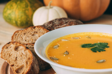 Vegetarian autumn pumpkin soup with rye homemade bread close-up. White bowl of soup garnished with parsley, pumpkin seeds and sesame seeds against a background of pumpkins.