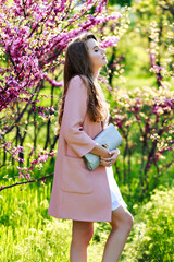 Attractive pretty young woman in light white dress, pink coat, with long hair walking in garden of spring time.