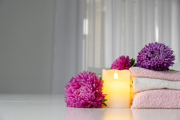 Set of fresh towels of pink and white color with purple aster flowers and candle on white table. Spa or massage salon concept. Copy space.
