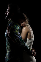 Couple posing on black background. Girl hugging boy in shadow from behind.