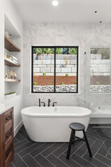 Gorgeous ensuite bathroom in luxury home. Features black herringbone tile floor, marble tile backsplash and wall,  freestanding soaker bathtub, and shelving.