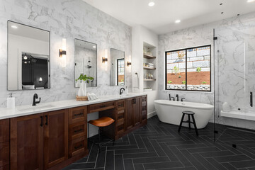 Gorgeous ensuite bathroom in luxury home. Features black herringbone tile floor, marble tile backsplash and wall,  freestanding soaker bathtub, double vanity and sconce lights.