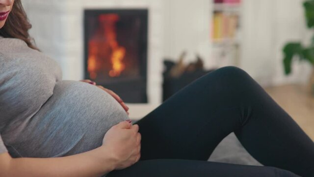 Beautiful pregnant woman with brown hair stitting in front of cosy fireplace stroking baby belly shot in 4k