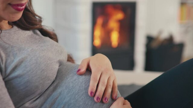 Beautiful pregnant woman with brown hair stitting in front of cosy fireplace stroking baby belly shot in 4k