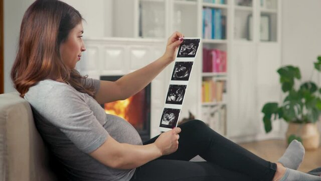Beautiful pregnant woman with brown hair stitting in front of cosy fireplace look at ultrasound pictures shot in 4k