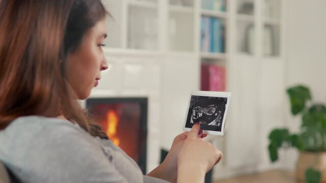 Beautiful pregnant woman with brown hair stitting in front of cosy fireplace stroking baby belly look at ultrasound pictures shot in 4k