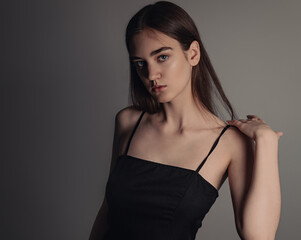 Fototapeta premium Beautiful woman with nude makeup and volume brown long hair looking mystery in black dress on grey background. Closeup. Art