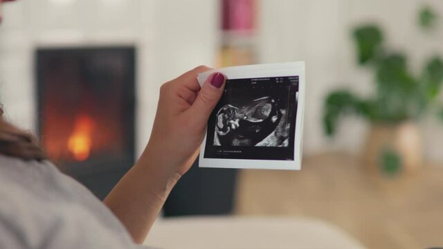 Beautiful pregnant woman with brown hair stitting in front of cosy fireplace stroking baby belly look at ultrasound pictures shot in 4k