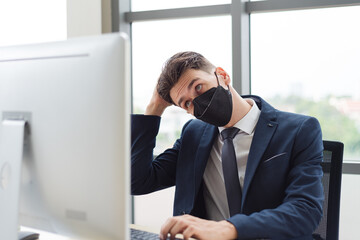 Adult smart business man in suit outfit wearing face mask sitting and working in front of computer typing keyboard with confusing emotion in modern office