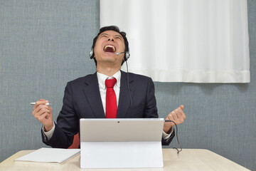 A young Asian man with a headset wearing business suit is in a trouble with customers and gets very angry during telework at home