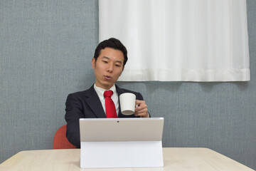 A young Asian man wearing business suit during telework at home takes a rest and drinks a cup of tea/coffee