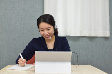 A young Asian woman with a headset wearing business casual clothes talks on a video meeting app and...