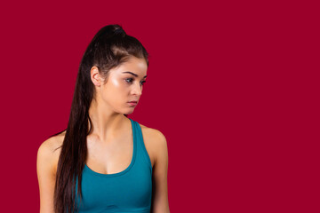 The profile photo of a sporty young girl in blue top and long brunette ponytail tired after training, poses over the red background in the studio.