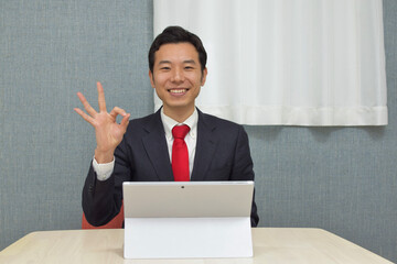 A young Asian man wearing business suit gets convinced and makes ok mark with his hand during telework at home