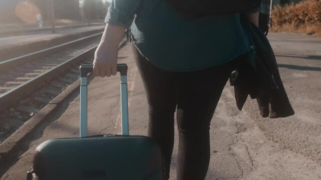 In sunny weather, a woman tourist with a suitcase walks slowly along the platform. Railroad tracks on the left