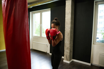 Fitness woman boxer in tight-fitting black sportswear and red boxing gloves training in a boxing...