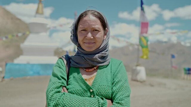 An outdoor shot of a middle aged  Asian woman with crossed arms is smiling and standing in the high altitude mountainous terrain against the Buddhist monastery looking at the camera 