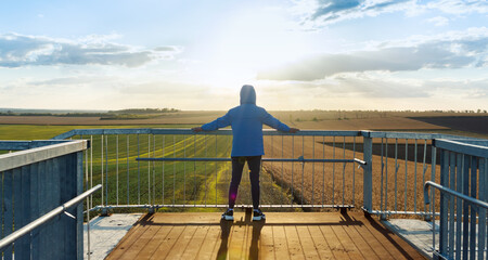 Young sports guy looks at the agricultural fields while standing on the bridge and lean on the railing. Evening time, rays of the setting sun, glare.