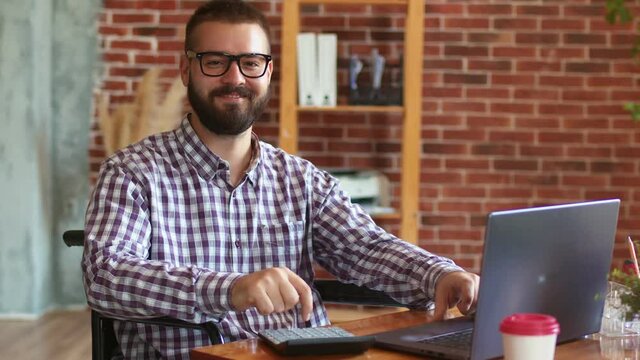 Disabled businessman sits on wheelchair at table with laptop, uses it, counts on calculator and looks ahead. Hipster businessman works remotely. Concept of remote work for people with disabilities