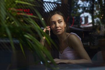 young asian pretty woman sitting at table of coffee shop, calling by smartphone. beautiful lady using cell phone shot through window glass outside of cafe. modern communication technology