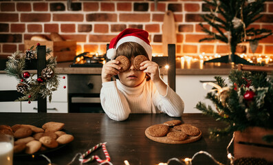 a little boy in a white knitted sweater and a New Year's hat is sitting at the table and closed his eyes with oatmeal cookies