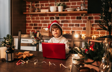 a Caucasian child in a white sweater and a New Year's hat is sitting at the kitchen table and typing a New Year's greeting on a laptop