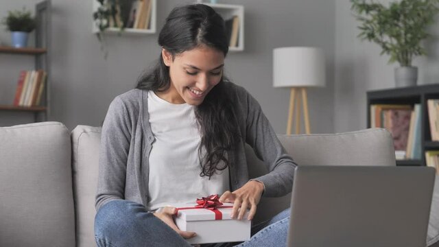Surprised young female opening gift box during a video call at the computer,happy arab latina woman sits on sofa opens present at the laptop,social distance online celebration