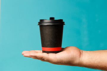 Mockup of man hand holding up a Coffee black color paper cup on blue background. Front view. Close-up of a hand. Concept coffee break. Coffee to go. Breakfast