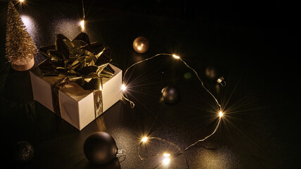Christmas composition. White gift box with golden ribbon, New Year balls and sparkling lights garland in xmas composition on black background for greeting card. Copy space.