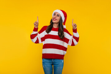 Fototapeta na wymiar Surprised happy beautiful Christmas woman in a red Santa hat and sweater is pointing up two hands while she isolated on yellow background