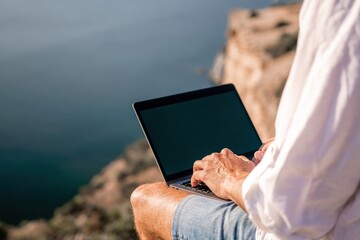 Digital nomad, man in the hat, a businessman with a laptop sits on the rocks by the sea during sunset, makes a business transaction online from a distance. Remote work on vacation.