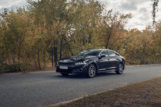 Samara, Russia - 09.12.2021: Black KIA K5 against the background of the autumn forest. The car is parked on an asphalt road near a colorful forest. Shooting a car at sunset. The front part