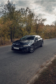 Samara, Russia - 09.12.2021: Black KIA K5 against the background of the autumn forest. The car is parked on an asphalt road near a colorful forest. Shooting a car at sunset. The front part