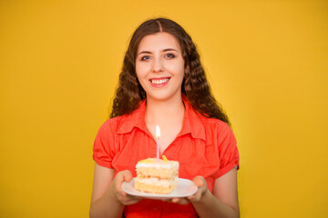 Portrait of a girl in a red shirt with a piece of cake and candle on a white plate in her hands isolated on a yellow background.