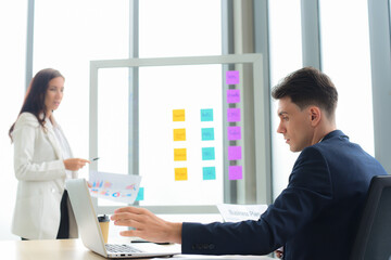 Caucasian male manager with laptop and listening to the out of focus female worker presentations of information and plans. presenting information on a glass board with sticky notes.