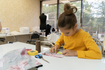 Cute little dressmaker working in atelier, handmade and handicraft. Kid in a sewing workshop examines pattern. Schoolgirl in the lesson draws a pattern of a soft toy