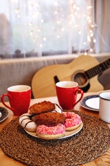 Tasty glazed doughnuts and two cups of hot beverage tea, coffee or cocoa on wooden table over window decorated with glittering glowing garland. Cozy winter season or christmas delicious sweets for two