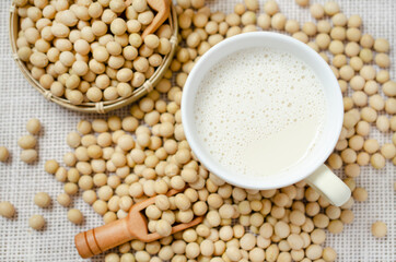 Top view of fresh delicious homemade soybean milk in soft yellow cup with dry soybean seeds on white sack fabric background