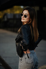 Fototapeta premium Young woman wearing black t-shirt, glasses and leather jacket posing against street