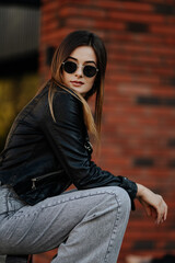 Young woman in sunglasses, hat and black leather jacket posing outdoor.