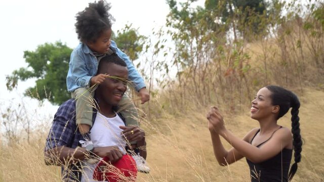 happy African American family in nature outdoors . black father Giving daughter Piggyback Rides On Walk in golden meadow . Parents carrying girl laugh on shoulders enjoying together at park . tease