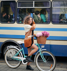 Fototapeta na wymiar Close up of smiling young woman with curly hair riding bicycle with bouquet pink fresh flowers on blue and white bus background. Concept of walking with bicycle in cetre city.