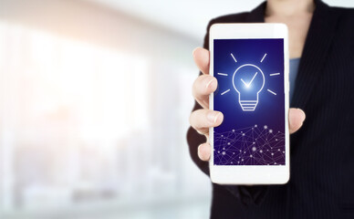 Creative idea.Concept of idea and innovation. Hand hold white smartphone with digital hologram light bulb sign on light blurred background. Business start up or goal to success.