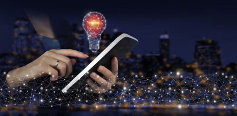 Network bright idea with light bulb. Hand touch white tablet with digital hologram light bulb sign on city dark blurred background. Ideas innovative technology and creativity.