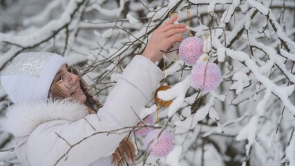 A girl hangs Christmas toys on trees in a snowy forest.