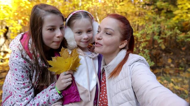 a cheerful woman takes selfies with her daughters in an autumn park, family photos of happy people