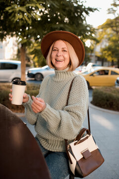 Beautiful Mature Woman 50-55 Year Old Drink Coffee In Paper Cup Wear Knit Sweater And Felt Hat  Sitting In Cafe Outdoors. Autumn Season. Healthy Aged Blonde Female. Happiness.