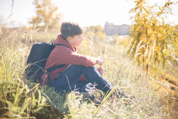 Naklejka na ściany i meble Autumn portrait of teenager boy sitting on grass. Young man with backpack resting relaxing, contemplating, thinking, daydreaming. Teen deep in thought Local travel. Active lifestyle People from behind