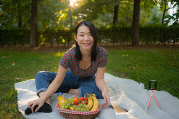 Autumn picnic at city park - lifestyle portrait of young happy and beautiful Asian Korean woman sitting grass with blanket and fruit basket enjoying relaxed a weekend picnic