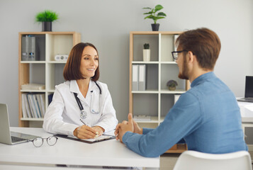Fototapeta premium Smiley female doctor in white lab coat with stethoscope talking to male patient in her office. Young man visiting modern clinic, hospital or medical center and discussing his treatment with physician